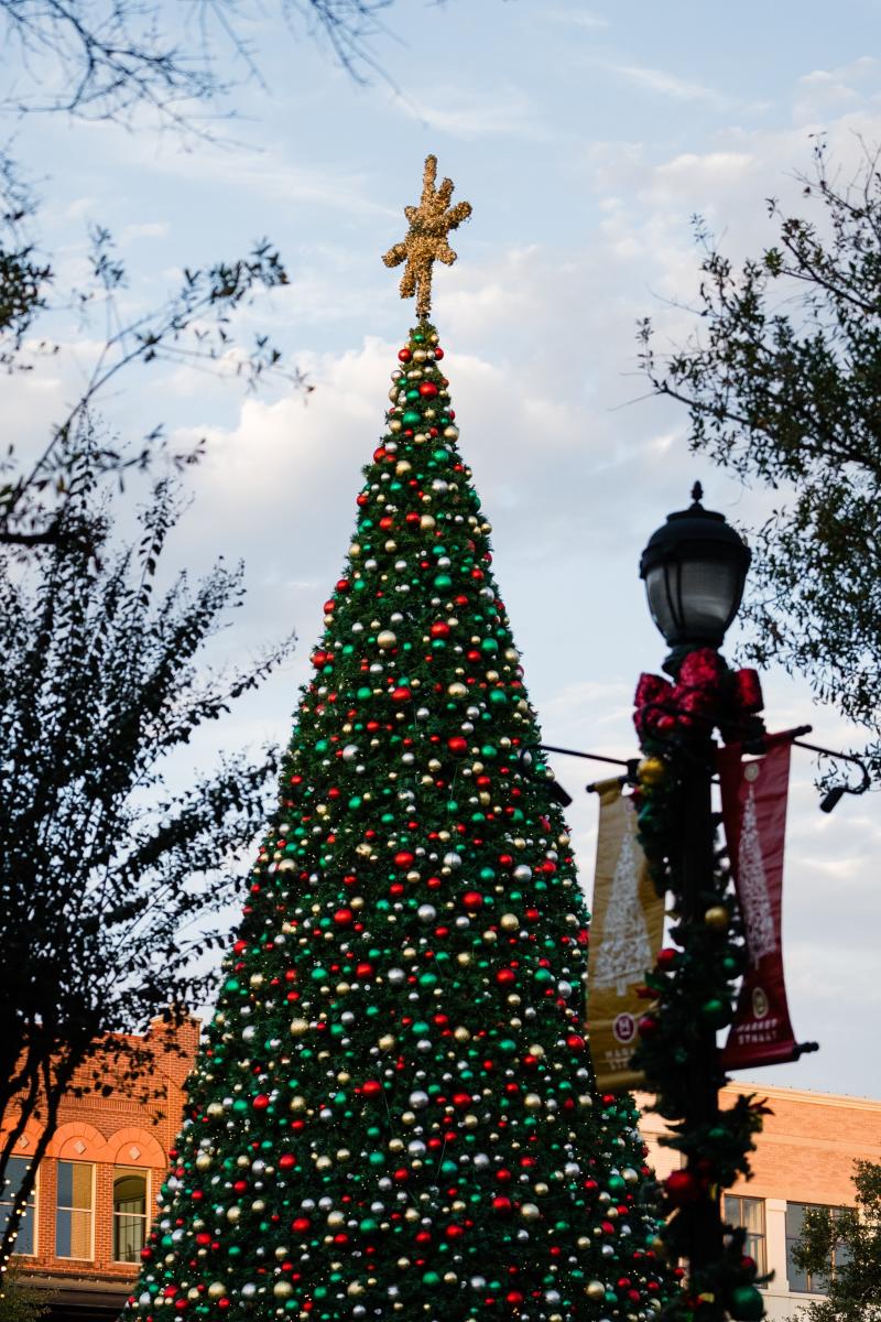 The Christmas Tree at Market Street is 70 feet tall, ringed with lights and decked with ornaments in red and green. A golden eight-point star rests at the top. Just in front of the camera, slightly out of focus, is a lamp post decorated with garland, red and green ornaments, and red and gold flags bearing Christmas trees and the Market Street logo.