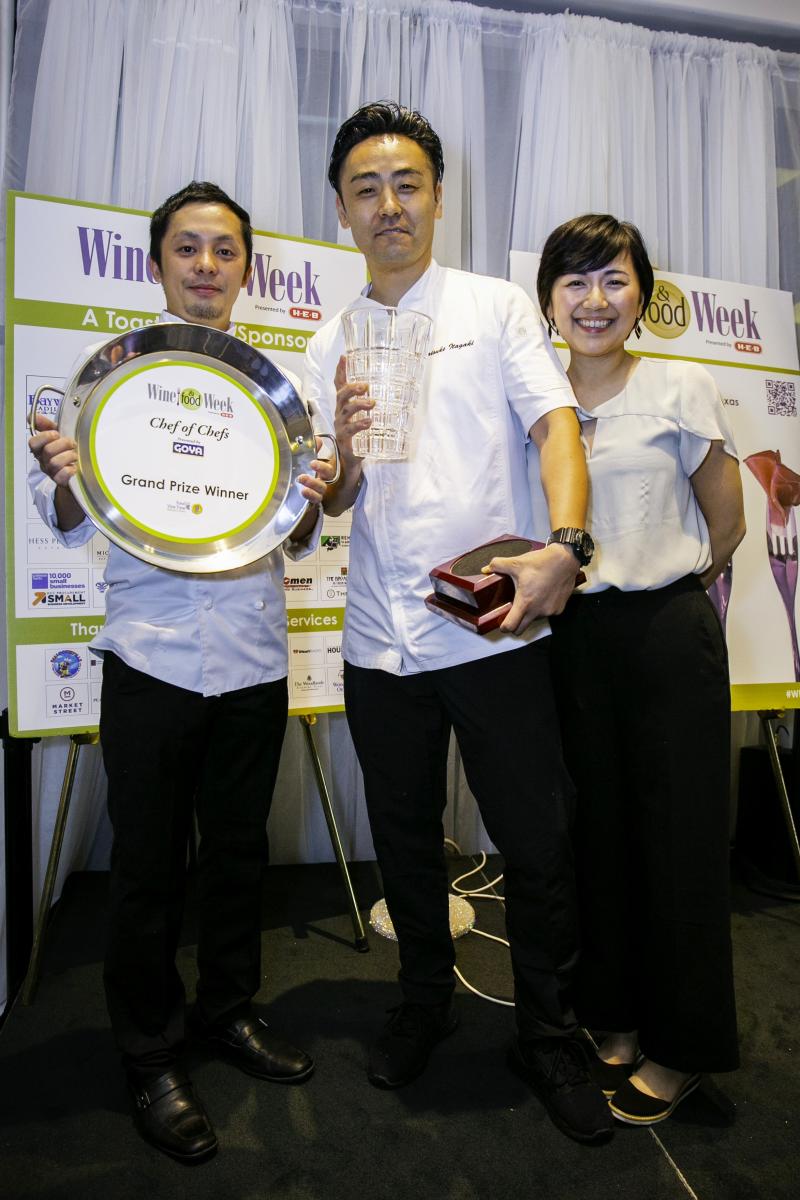 Three young people (two men, one woman, all of Asian descent) smile at the camera. The men are dressed in white chef uniforms, and the woman is wearing a white blouse and black slacks. The man on the left is holding a metal pan that reads, "Wine & Food Week | Chef of Chefs, Presented by GOYA | Grand Prize Winner." The man in the middle is holding a glass vase (the trophy) and a solid maroon trophy stand. Behind them is a sheer white curtain and miscellaneous posters for Wine & Food Week.