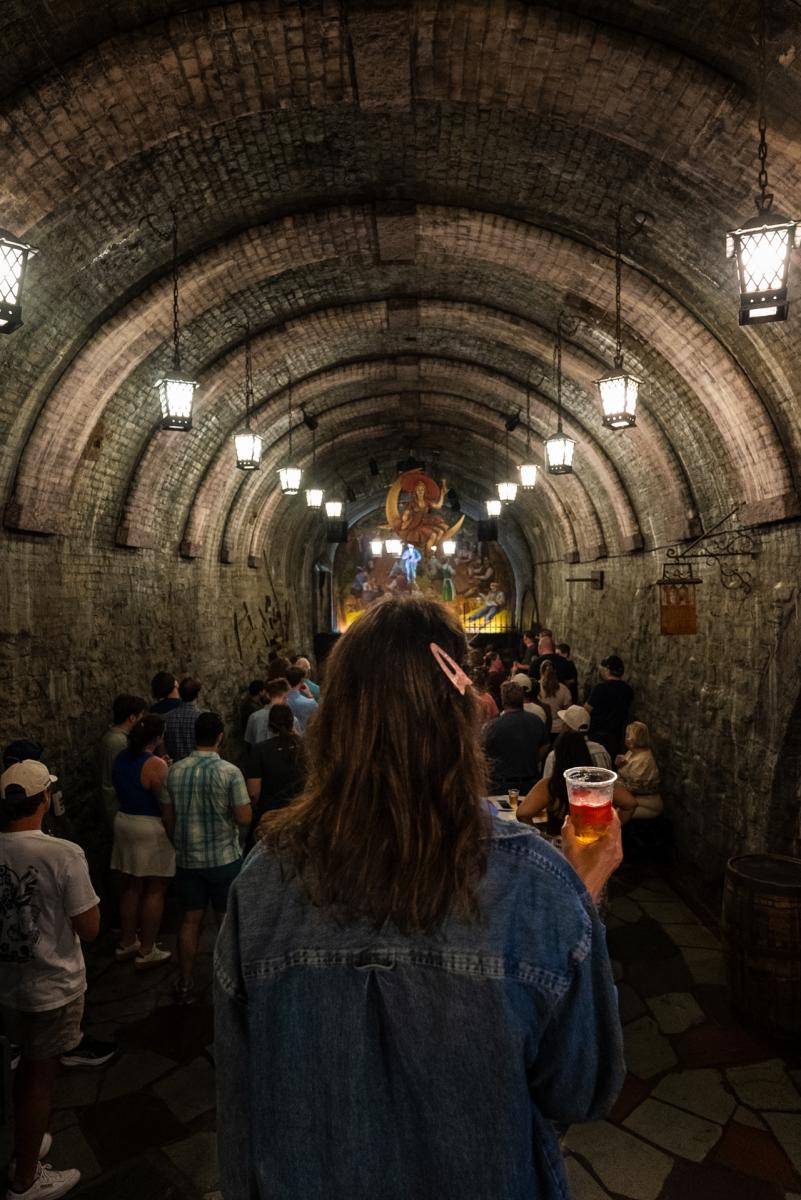 A visitor holding a beer follows a tour group through the historic Miller Brewery caves in Milwaukee, beneath arched brick ceilings and hanging lanterns.