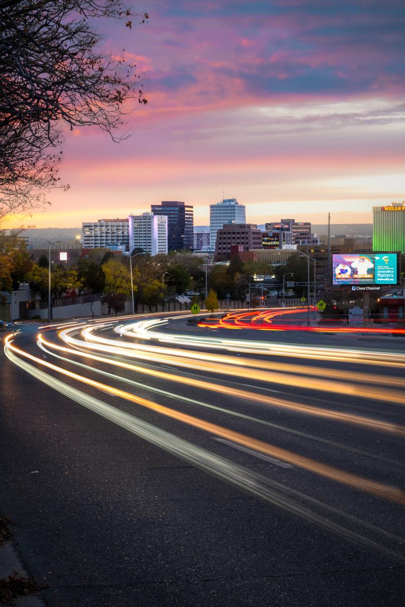 A city skyline at dusk with a road in the foreground showing light trails from passing vehicles. The sky is painted in bright colors during a sunset. Several tall buildings are visible, including recognizable structures with billboard advertisements. Trees with sparse leaves line the road
