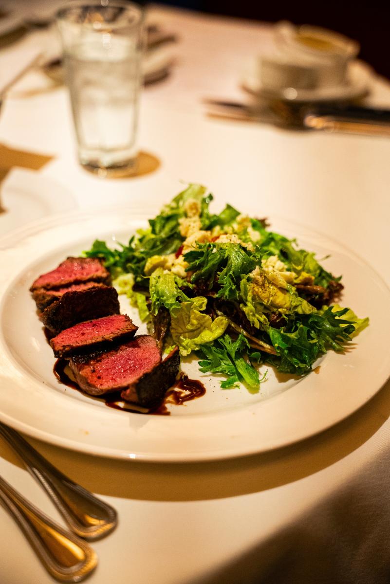 The camera focuses on a small cobb salad with pink tenderloin slices. A solid white plate, tablecloth, silverware, and a glass of water are out of focus to better highlight the food.