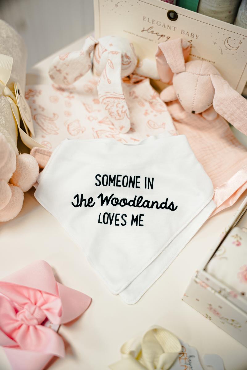 A white baby bib that reads (in black text), "SOMEONE IN ~The Woodlands~ LOVES ME." Around it are various pale pink and white baby bows and toys.