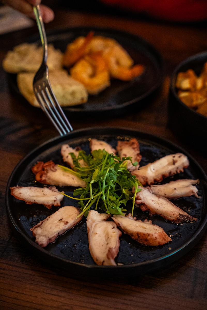 The camera focuses on slices of octopus placed in a circle around a green garnish. A fork reaches in to pick one up. Behind it, out of focus, is a plate of shrimp and rolls.