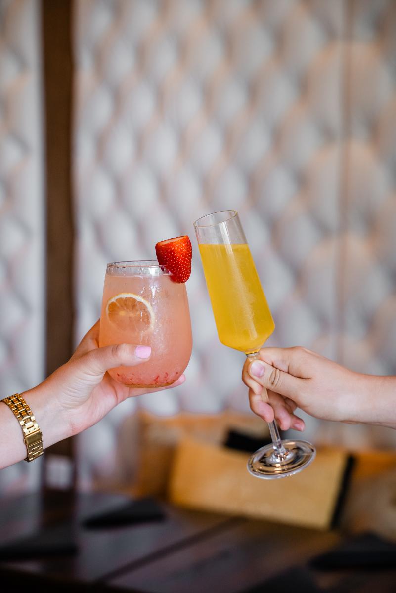 Two women cheers their glasses at La Rambla. On the left, a woman holds a round, squatty glass of a pink drink with a strawberry on the rim and an orange slice floating within. On the right, a woman holds a tall, skinny glass with a sunny yellow mimosa. Behind them is a white padded booth and dark wood table.