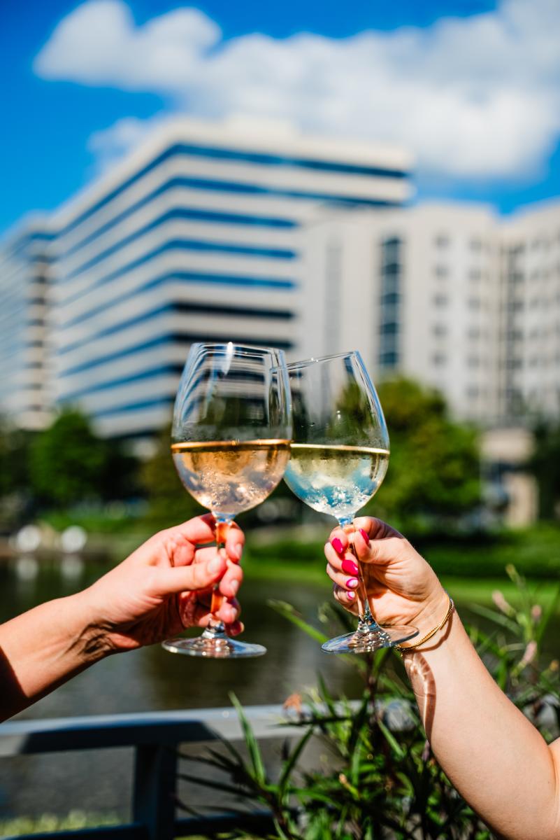 Two women (off-screen) raise their glasses in a toast. The glass on the left is half-filled with Rose, the other with white wine. The woman on the right is wearing a gold bracelet and bright pink acrylic nails. In the background is Lake Woodlands, lush greenery, and office buildings with wraparound windows reflecting a bright blue sky.