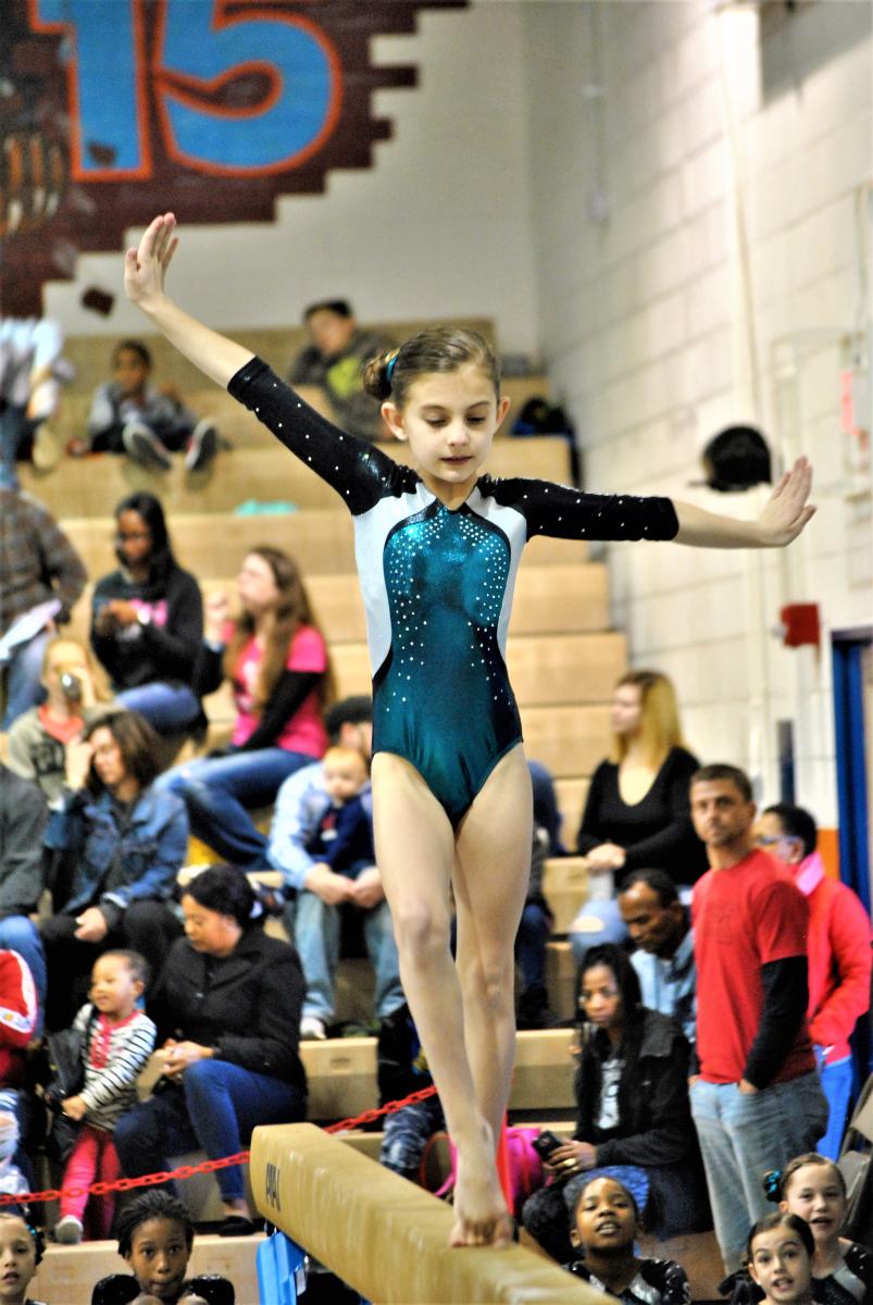Young gymnast in a teal and black leotard balances on a beam with arms extended while spectators watch from the stands in the background