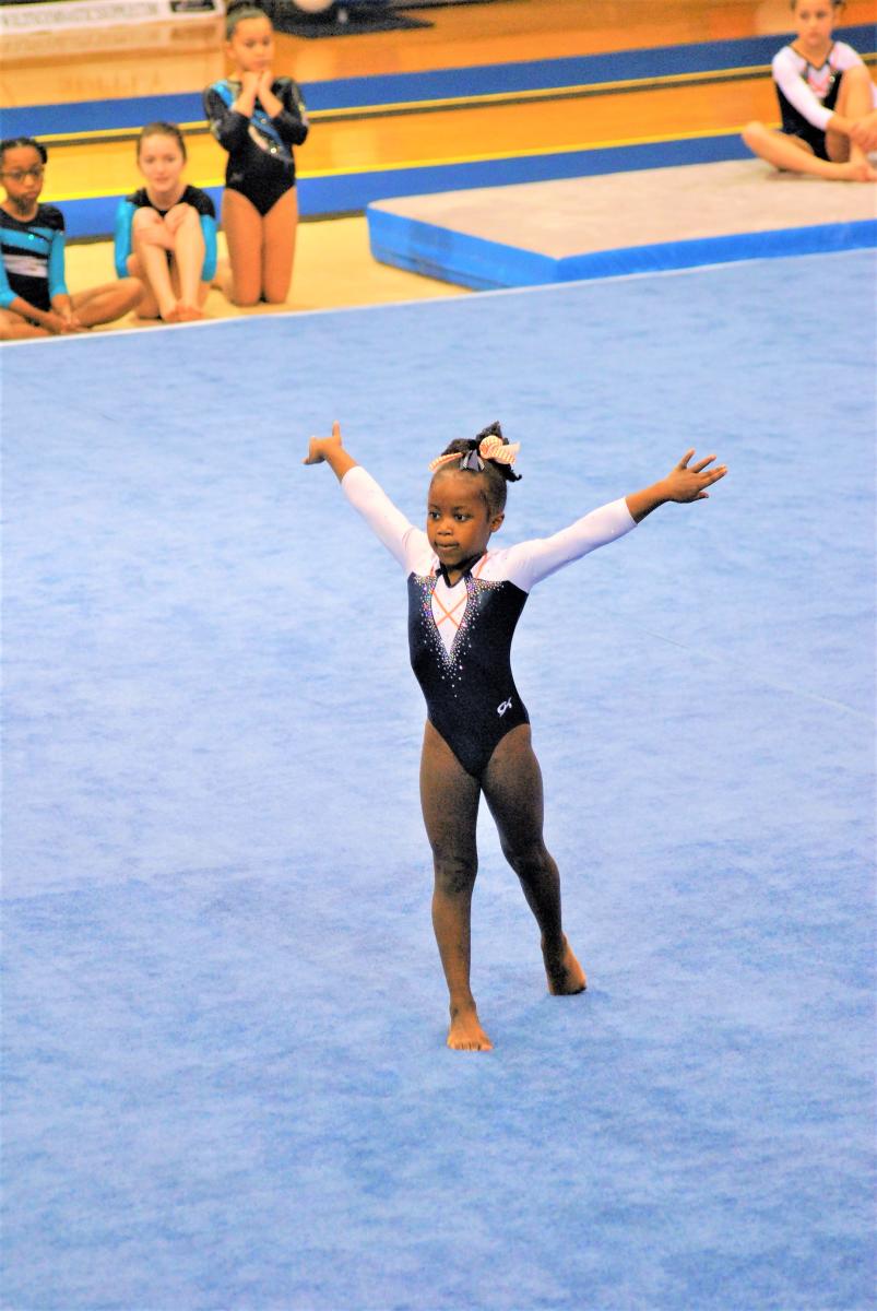 Young gymnast in a black and white leotard performs a floor routine with arms extended while other gymnasts watch from the mat in the background