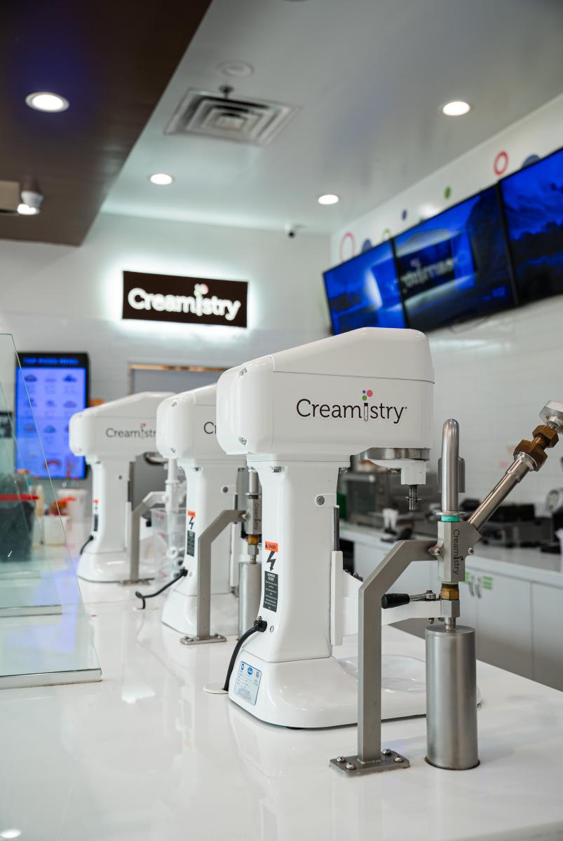 The camera peers behind the glass shield along the counter, focused on three stand-up mixers bearing the Creamistry name/logo. In the background (out of focus) is a larger neon sign bearing the shop name, an electronic menu, and a row of TVs.