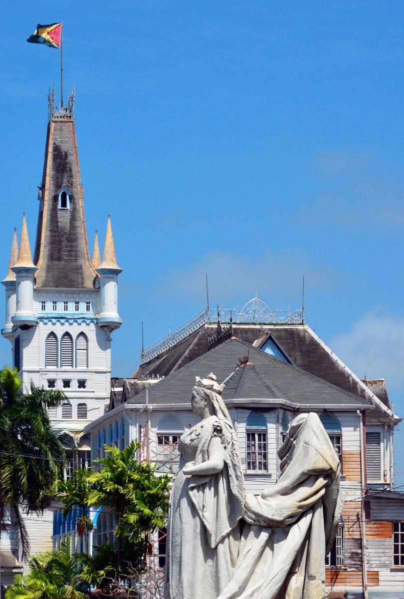 A historical building with a tall, Gothic-style tower and a statue of Queen Victoria in front  of it. The building has a blue and white facade and a sloped roof. A flag of Guyana is flying at the top of the tower.