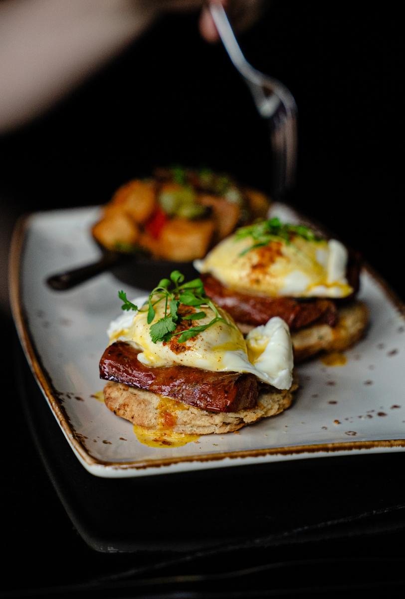 The camera focuses on a ceramic plate of two eggs benedicts with bison sausage and poached eggs on a flaky biscuit. Behind them, though out of focus, is a handheld cast iron skillet of seasoned potatoes. The client brings a fork over the potatoes to eat.