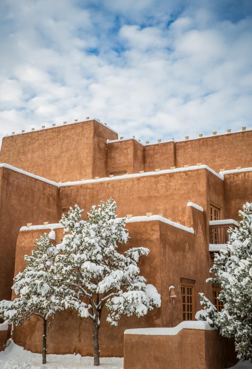 Snow-covered trees in front of the adobe architecture under a blue sky with fluffy clouds.