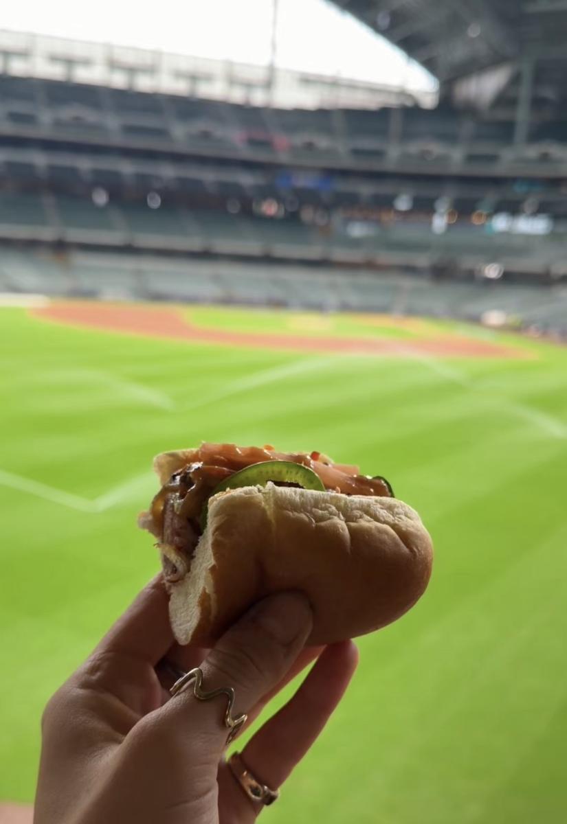Hand holding a ballpark sandwich topped with meat and jalapeño in front of the field at American Family Field.