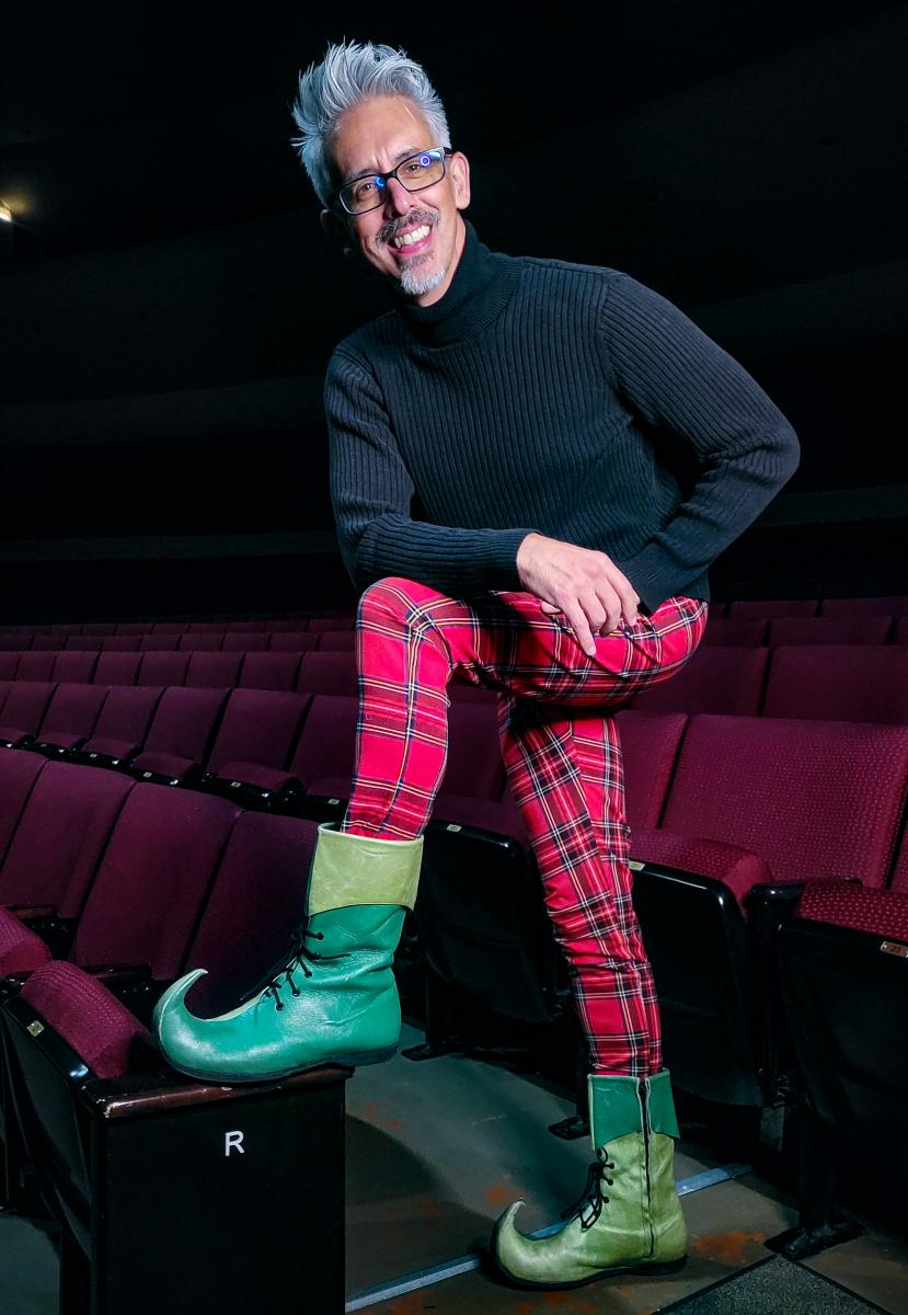 Brian Marcum, artistic director of Music Theatre Wichita, poses in plaid pants and elf shoes in a dim theatre