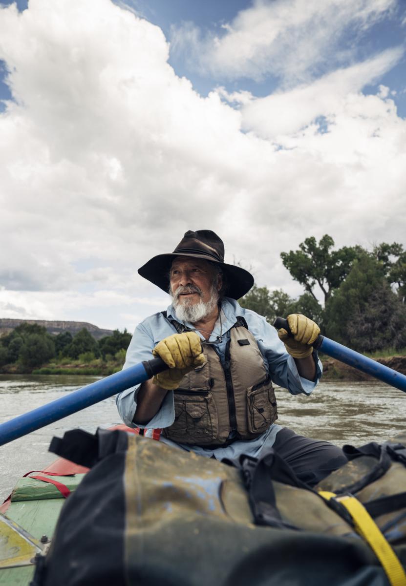 Person rowing a raft down a river, wearing a life jacket and a wide-brimmed hat, with scenic trees and cloudy sky in the background.