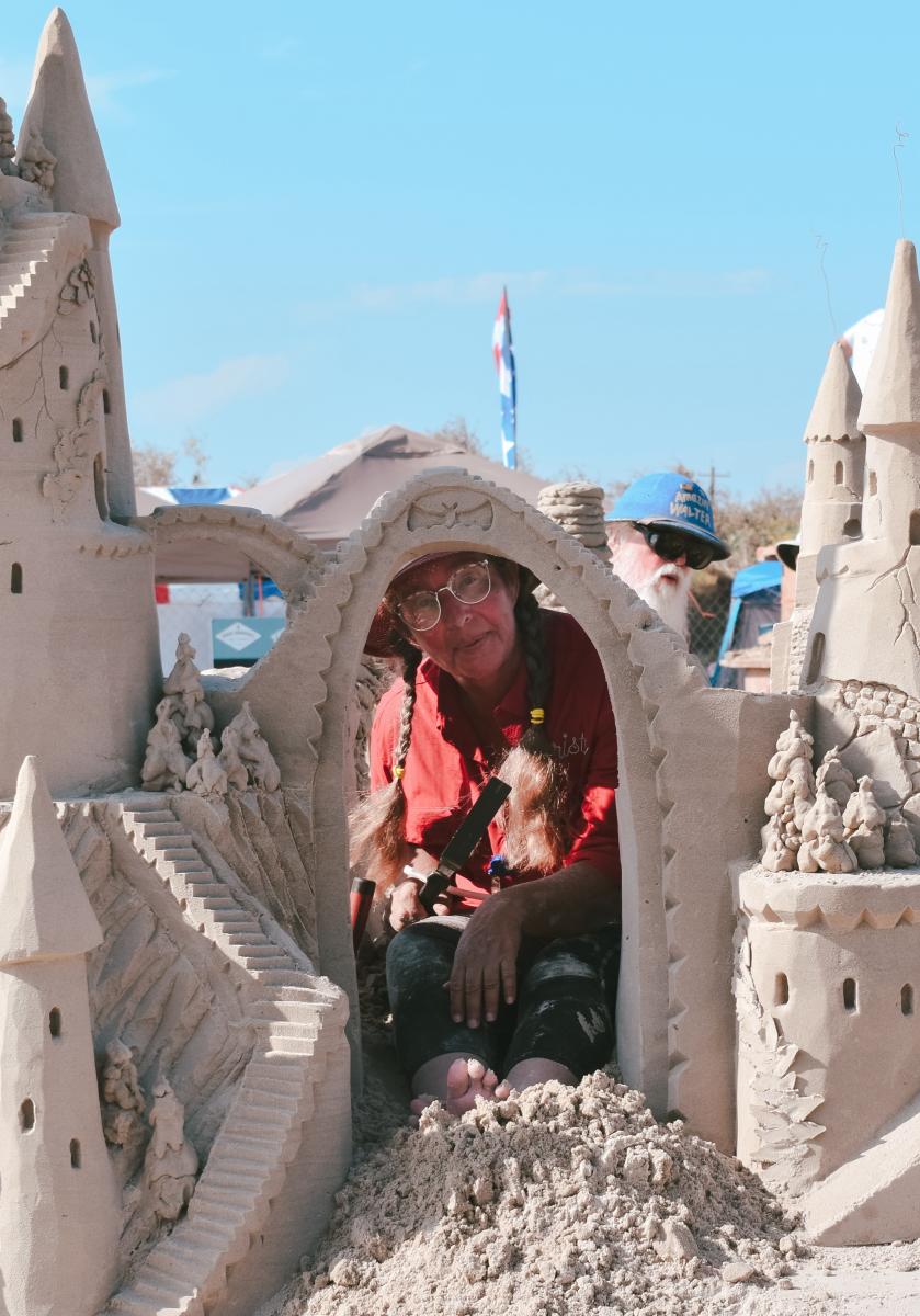A woman in braids smiles a the camera through the arch of a sandcastle she is working on sculpting.