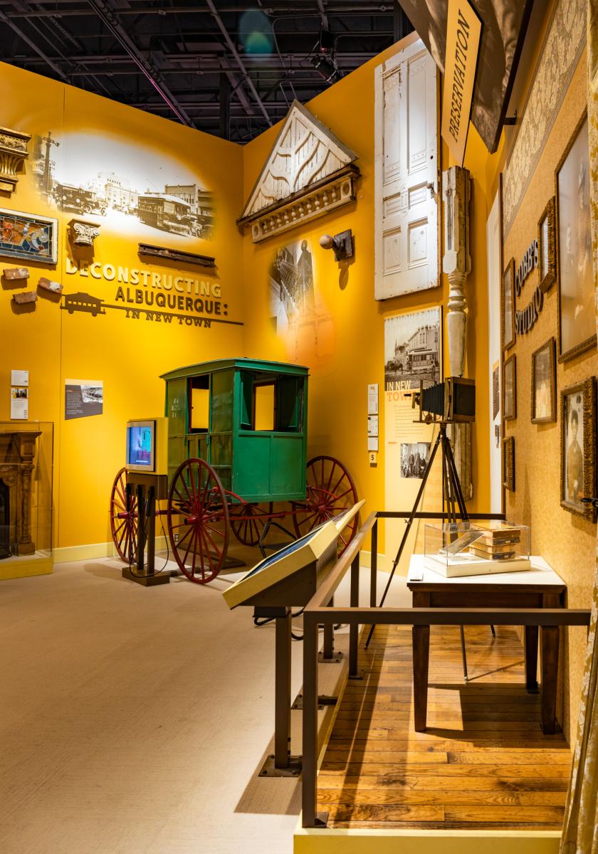 Interior view of the "Reconstructing Albuquerque" exhibit, featuring a bright yellow wall with historical documents on display, an old-fashioned green and red carriage, and an antique surveyor's transit.