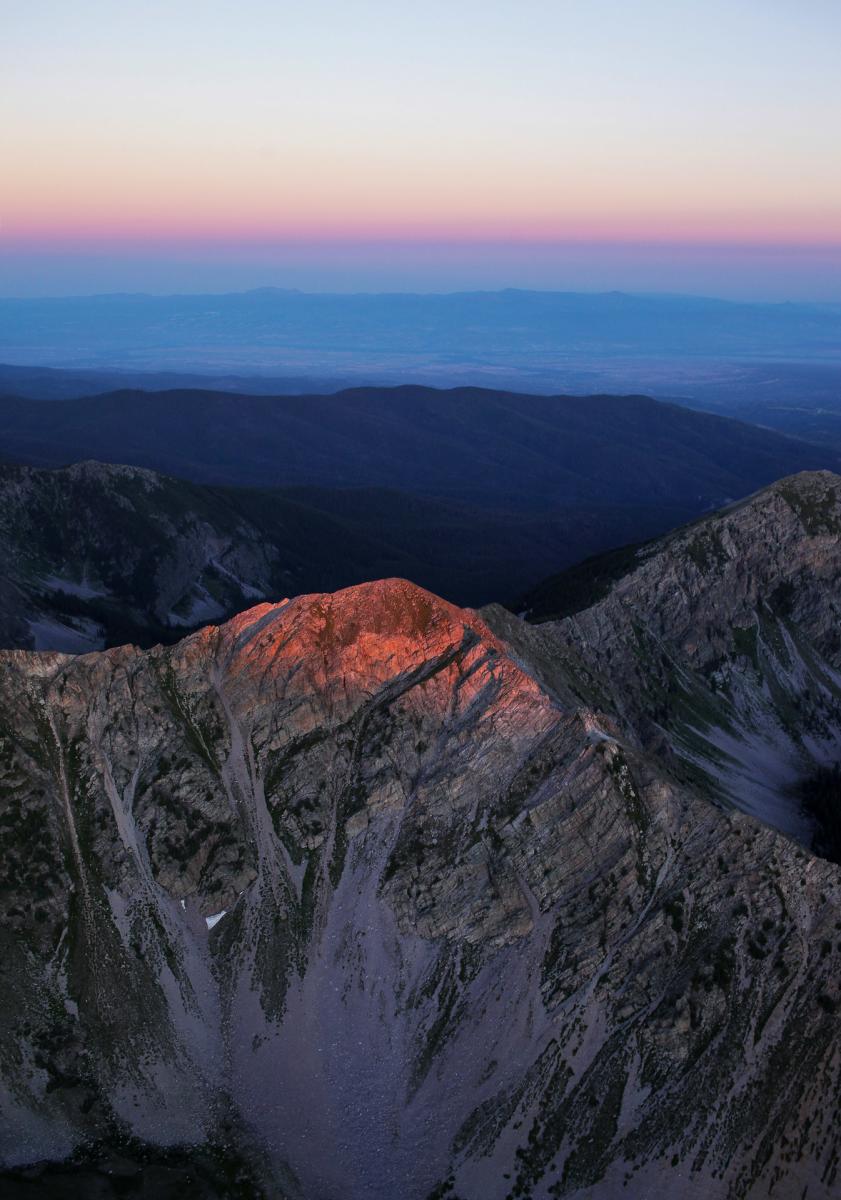 A mountainous landscape at dusk, with the peaks bathed in a warm light. The view includes distant ranges under a gradient sky.