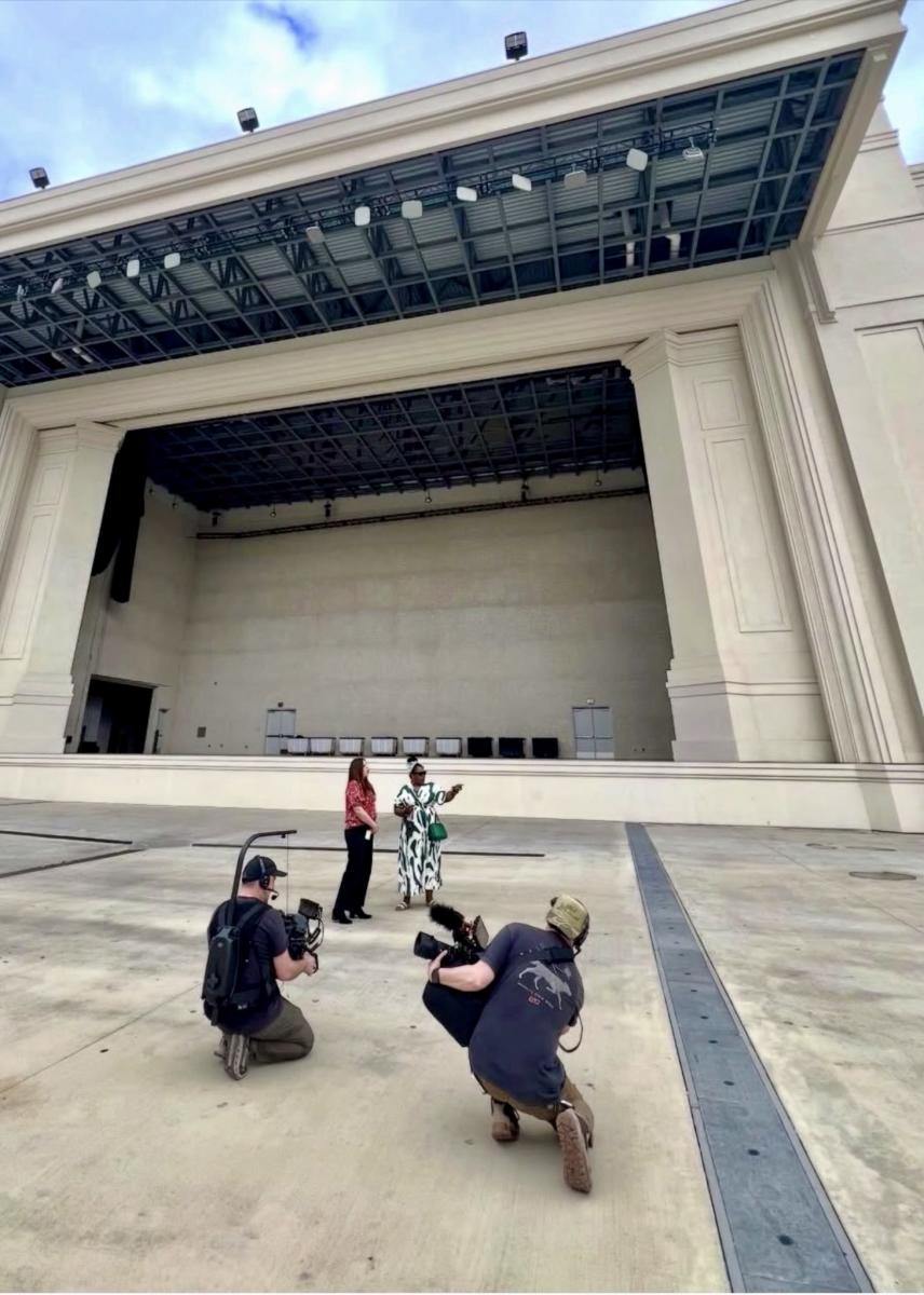 Leslie Robinson is standing with another lady in front of the Orion Amphitheater while two cameramen are filming her visit.