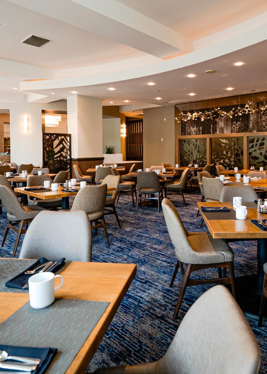 The camera overlooks the new Acqua Restaurant from the back corner of the room. The space features at least twelve tables that each seat 2-4 guests. Each table is set with a modern gray placemat, navy cloth napkins, silverware, plain white coffee mugs, as well as salt, pepper, and sugar. The room dividers are dark solid wood frames with black metal inlays depicting tropical plant fronds. Along the entryway is a string of decorative glass and metal lights.