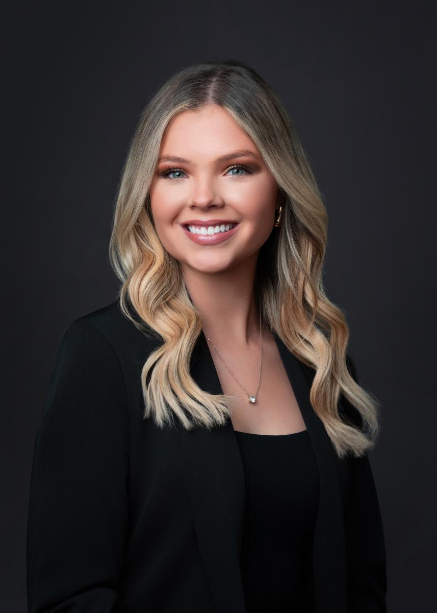 A young woman smiles at the camera. Her blonde hair is curled loosely over her shoulders. She is wearing a small silver pendant, a black top, and a black sweater on a black backdrop.