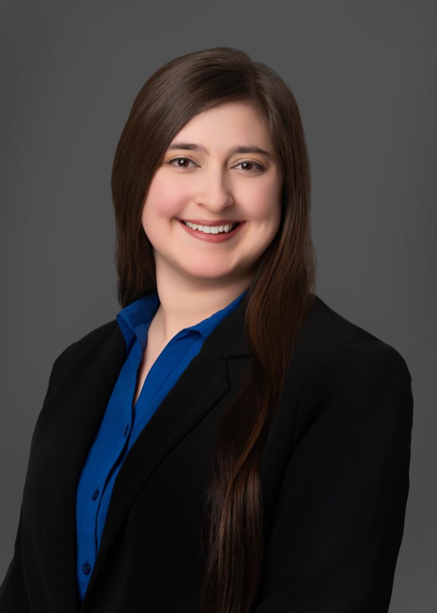 A young white woman with long brown hair smiles at the camera. She is wearing a bright blue button-down shirt and a black blazer in front of a solid gray background.