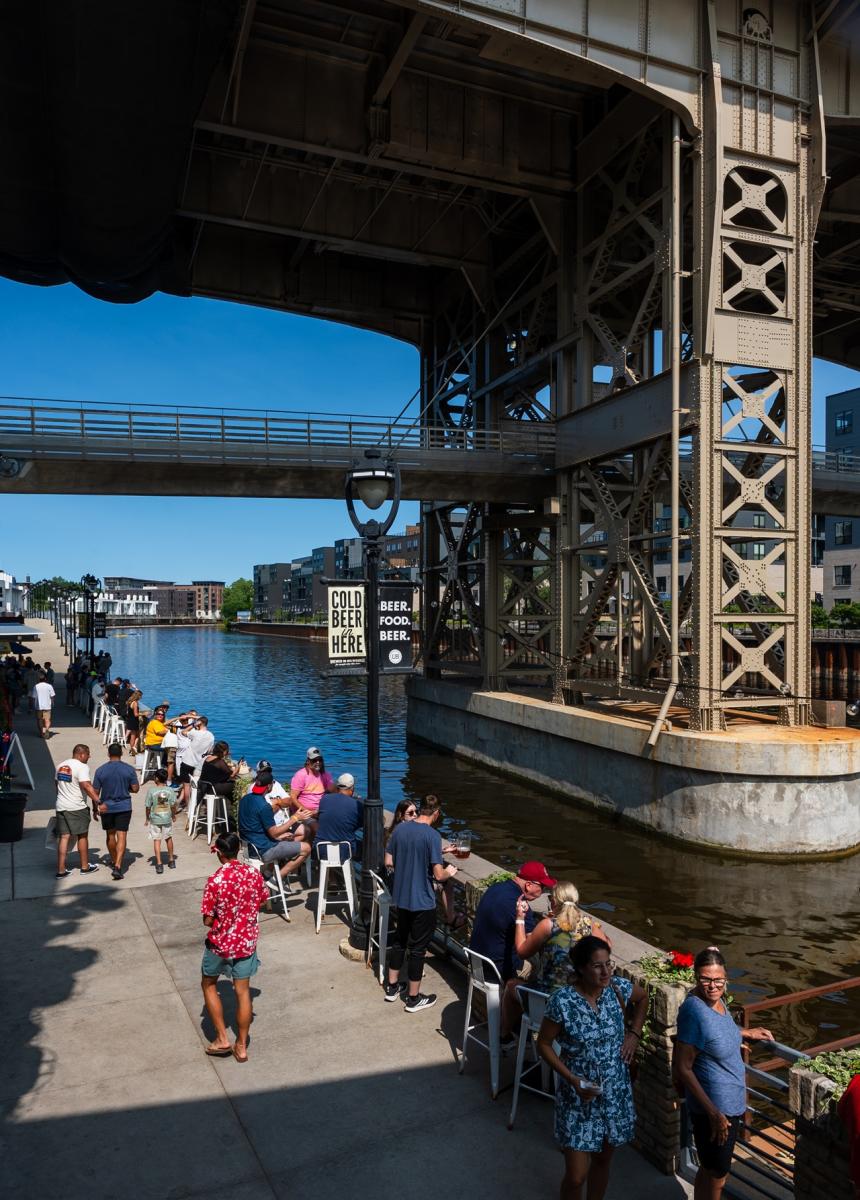 Visitors enjoy drinks and outdoor seating along the Milwaukee River outside Lakefront Brewery, beneath the towering Holton Street Bridge on a sunny day.