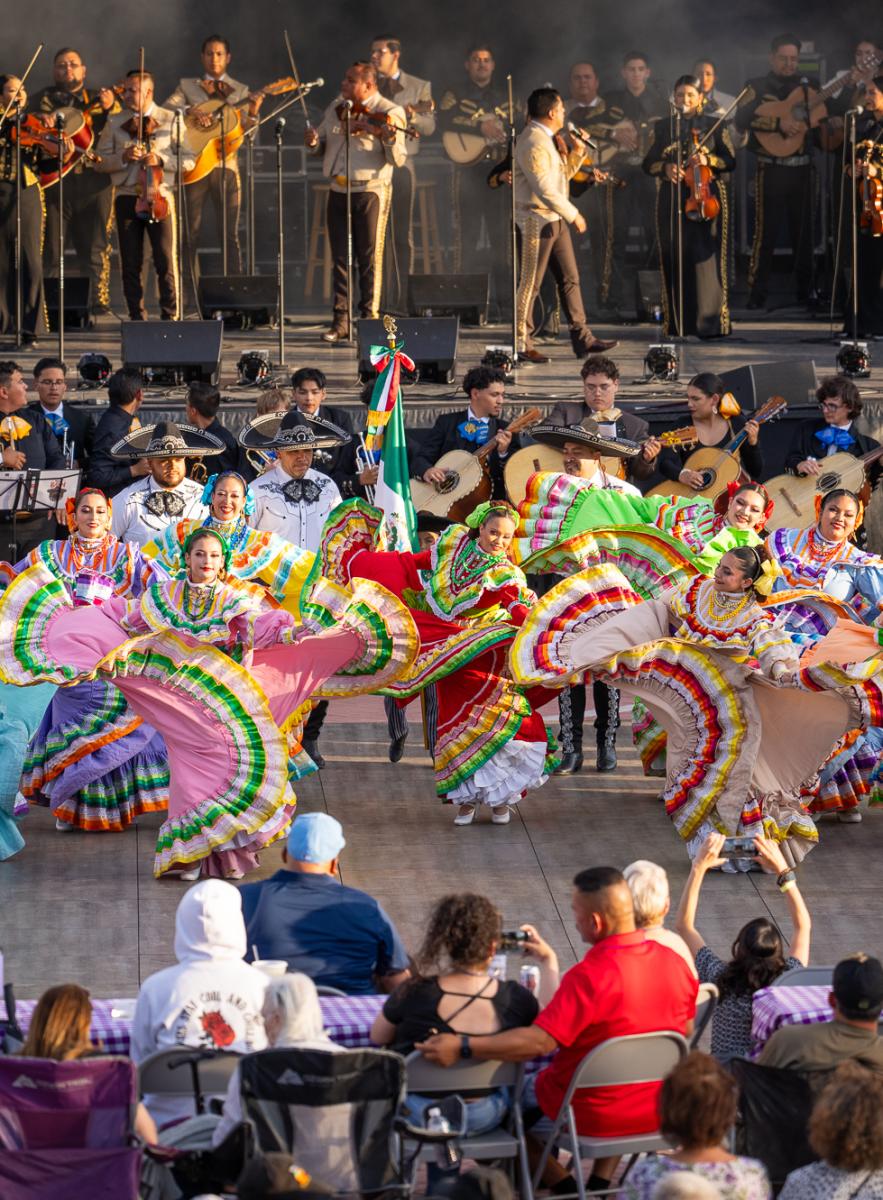 Performers in vibrant, traditional Mexican costumes dance at a folkloric festival, accompanied by a large mariachi band on stage, with an audience in the foreground.