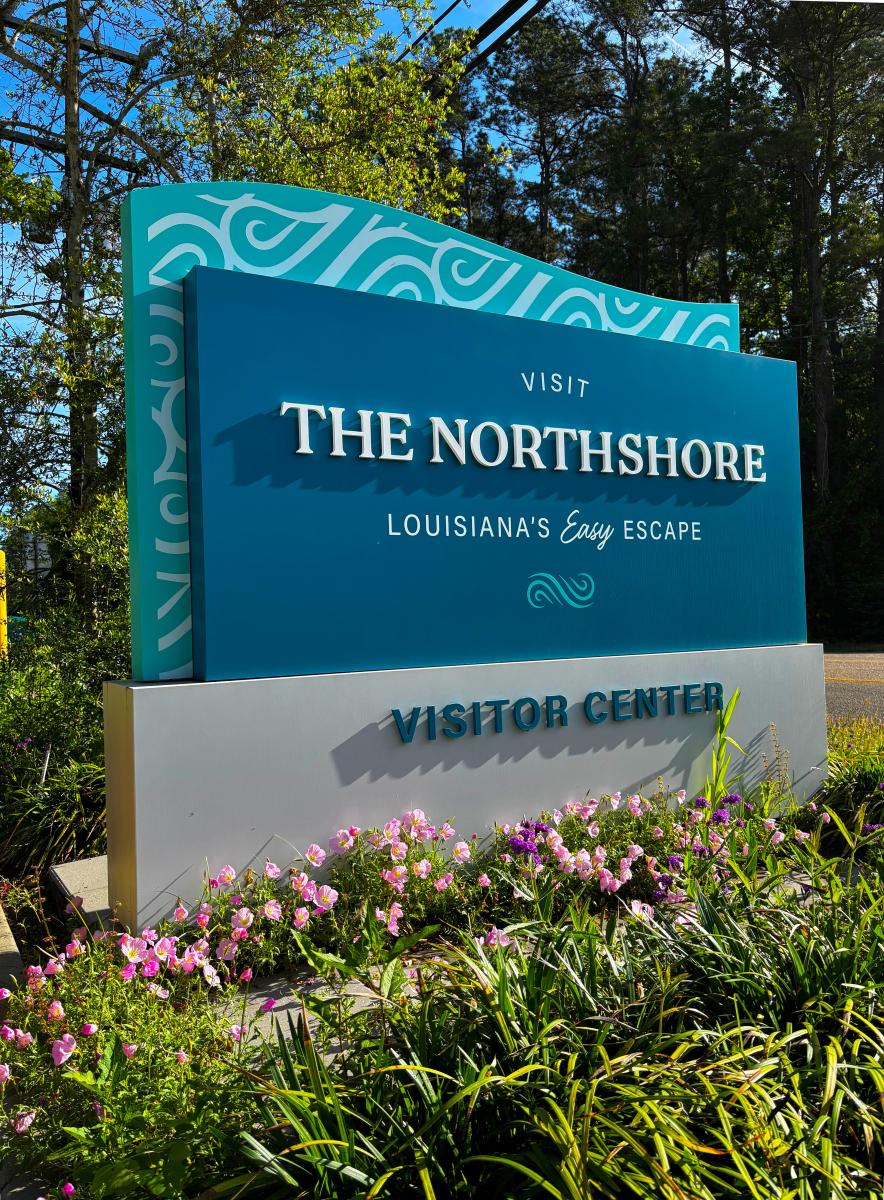 Visit The Northshore Visitor Center sign beside Highway 59 in Mandeville, Louisiana, surrounded by blooming flowers and landscaped greenery.