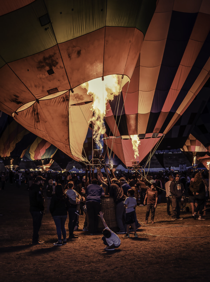 Crowd gathers around illuminated hot air balloons at night, with flames glowing warmly.