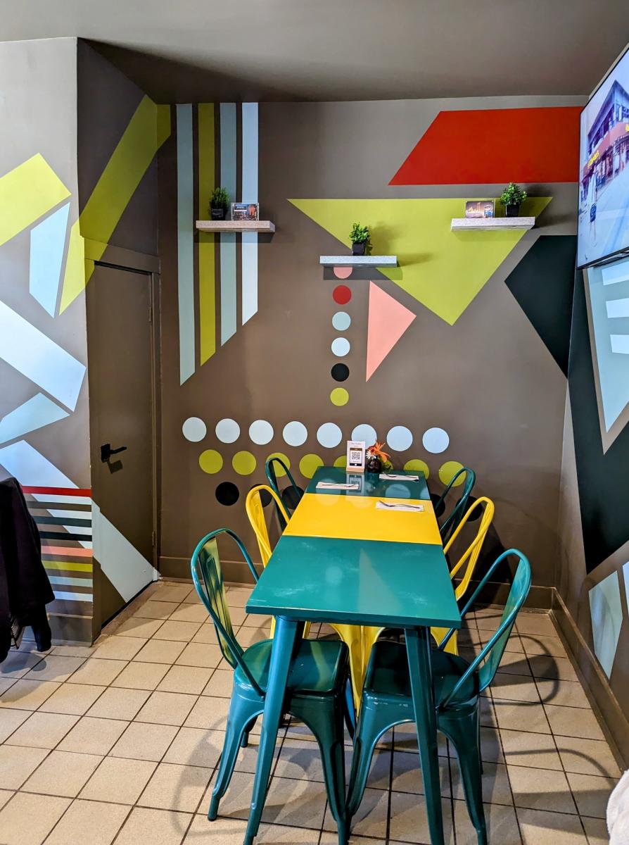 Image is of colorful table and chairs with geometric designs and colors on the wall. Also hanging shelves with plants.
