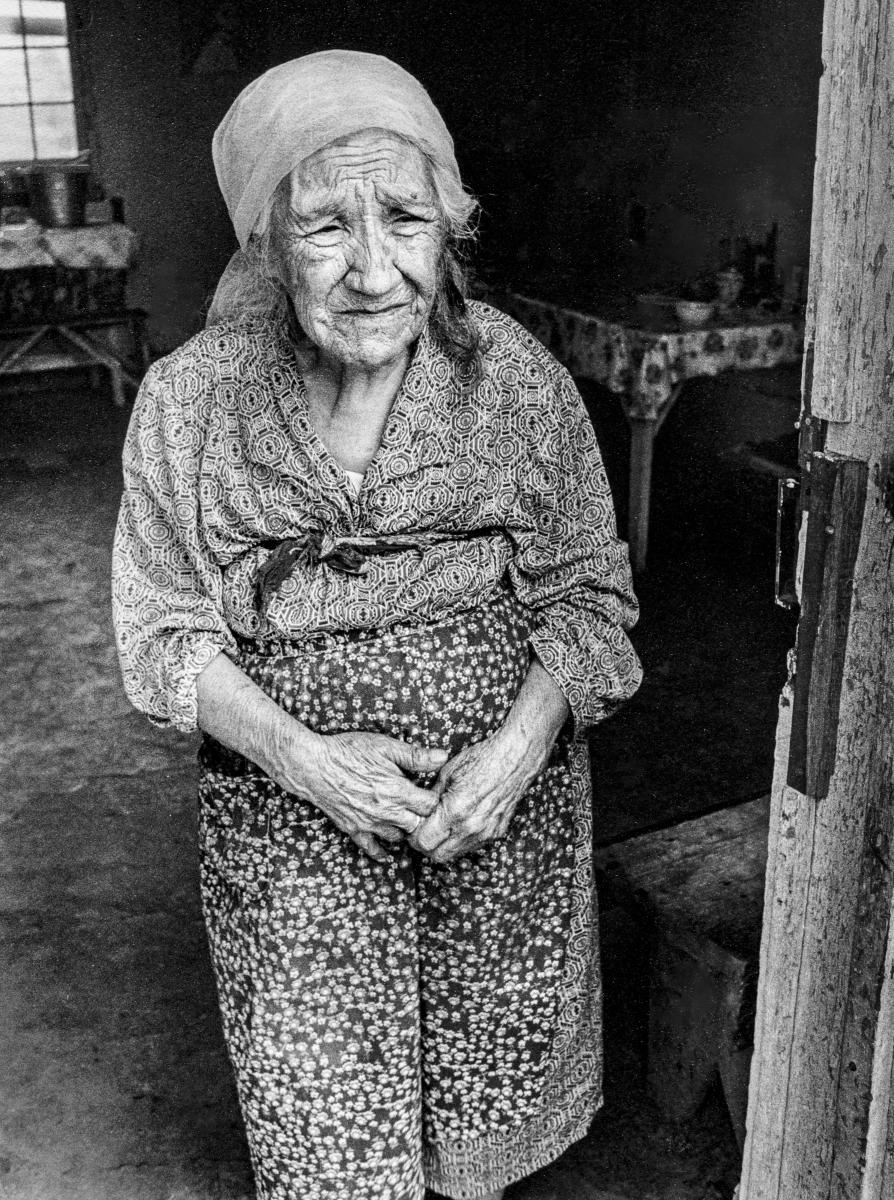 Black and white portrait of an elderly woman standing in the doorway of her home, wearing a patterned dress and head covering.