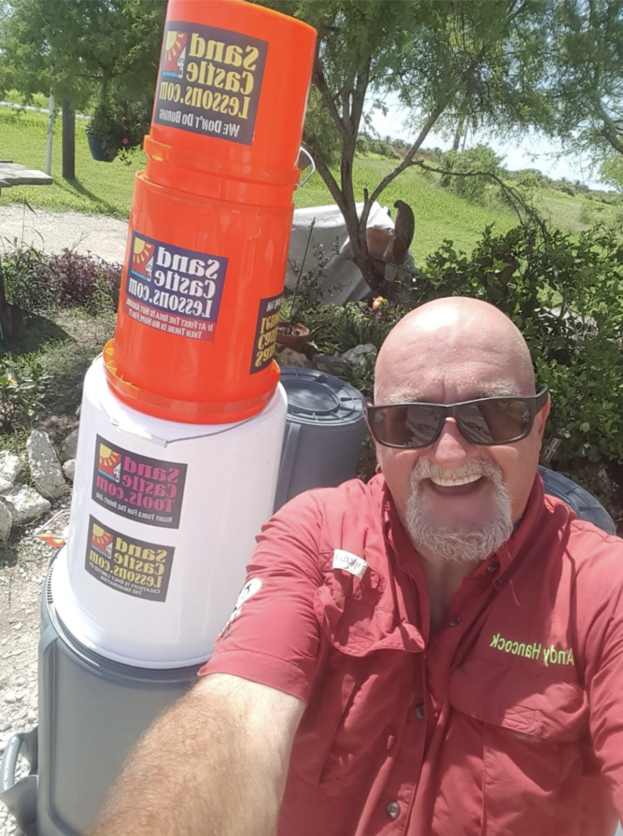 Selfie taken by a bald man in a red button down with sunglasses on. He smiles at the camera as he stands next to a tower of five gallon buckets.