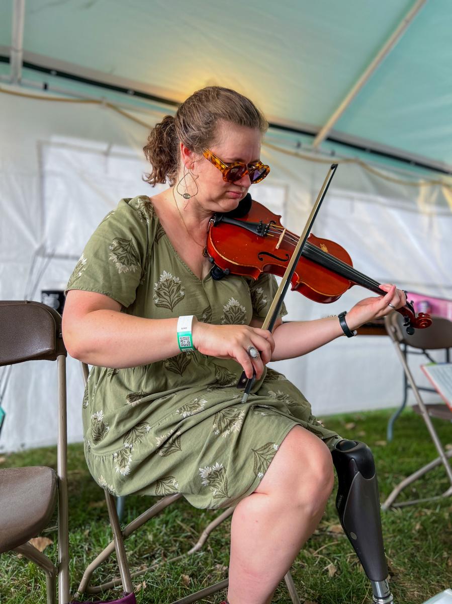 A musician seated on a chair under a tent, playing a violin. They are wearing a green dress and sunglasses, and have a festival wristband on.