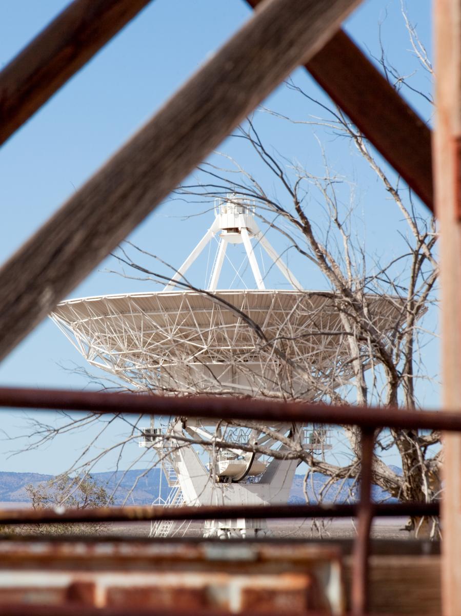 A large satellite dish viewed through a geometric frame of crisscrossing wooden beams, set against a clear blue sky and distant mountains.