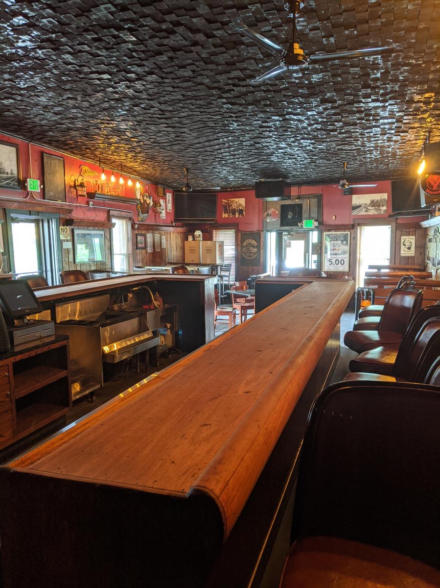 An interior of a bar. A wooden bar crosses the photo with seats along it. The ceiling is black and textured.