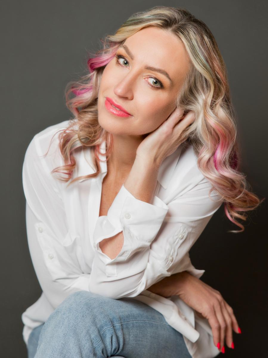 A headshot of a blonde woman wearing a white button-down shirt and light blue jeans. Her professionally curled hair is ash blonde with lighter blonde and hot pink highlights. Her lipstick and nails are also hot pink. She is posing with her elbow resting on her knee and her left hand lightly supporting her head.