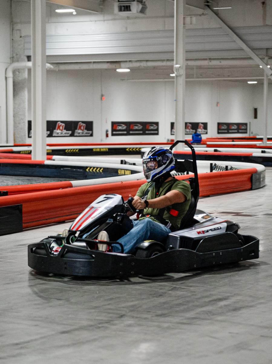 A man turns a go kart around a corner at K1 Speed. He's wearing a green T-shirt, blue jeans, and a black, white, and blue motorcycle helmet. The track is gray and rubbery, the barriers alternating in red and white stripes. K1 posters line the back wall.