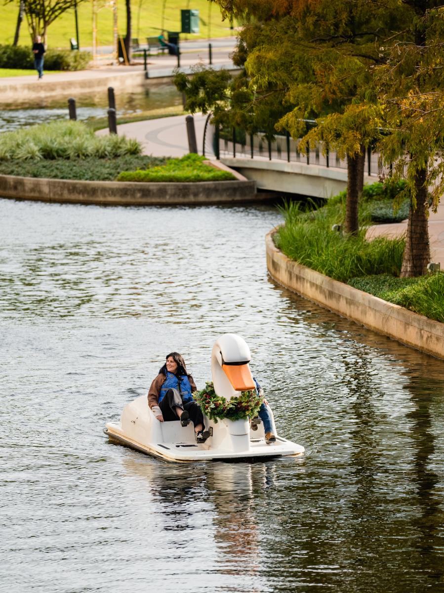 A couple paddles a swan boat down The Waterway (toward the camera). Only the woman is visible; she has long dark hair, a brown shirt, and black pants. She's also wearing a bright blue life jacket. The scenery behind them features a footbridge and fall foliage.