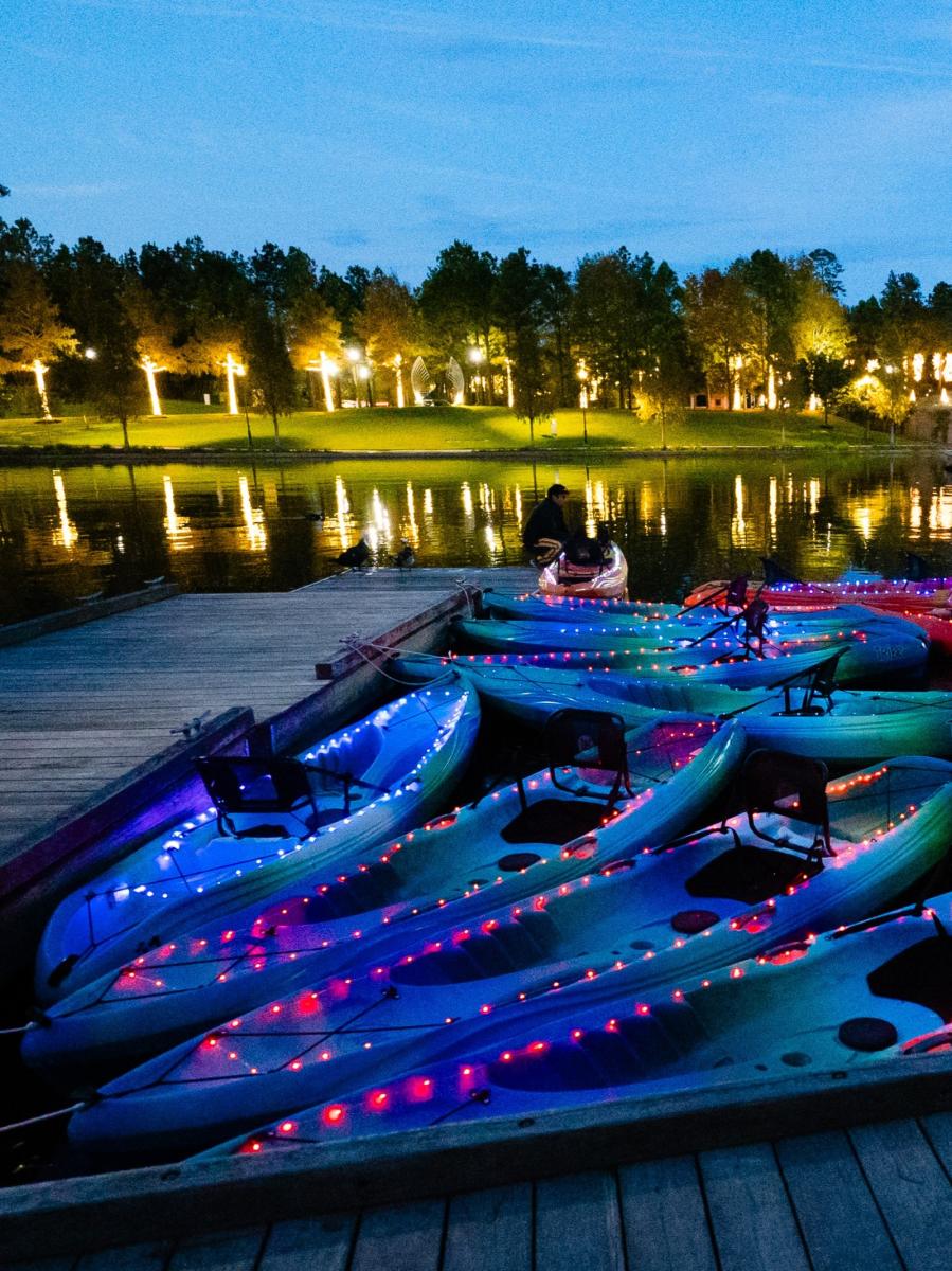 Brightly colored kayaks (some blue and purple, others pink, orange, and yellow) rest in the water by the docks at Riva Row Boat House. Each kayak is lined with a string of colorful Christmas lights for the Moonlight Paddle.