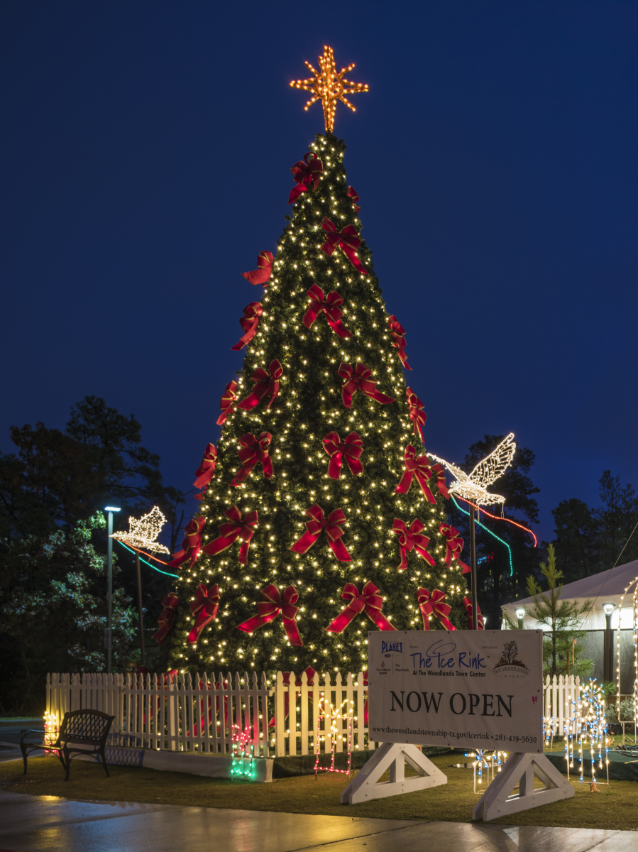 The Christmas Tree outside The Woodlands Ice Rink is decked with golden lights and large red ribbons. Two light displays shaped like doves take flight on either side of the tree. A white picket fence surrounds the tree, and beside it is a sign that reads, "The Ice Rink at The Woodlands Town Center | NOW OPEN."