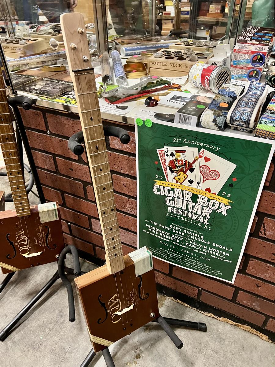 Two handmade cigar box guitars on stands next to a green cigar box festival flyer.