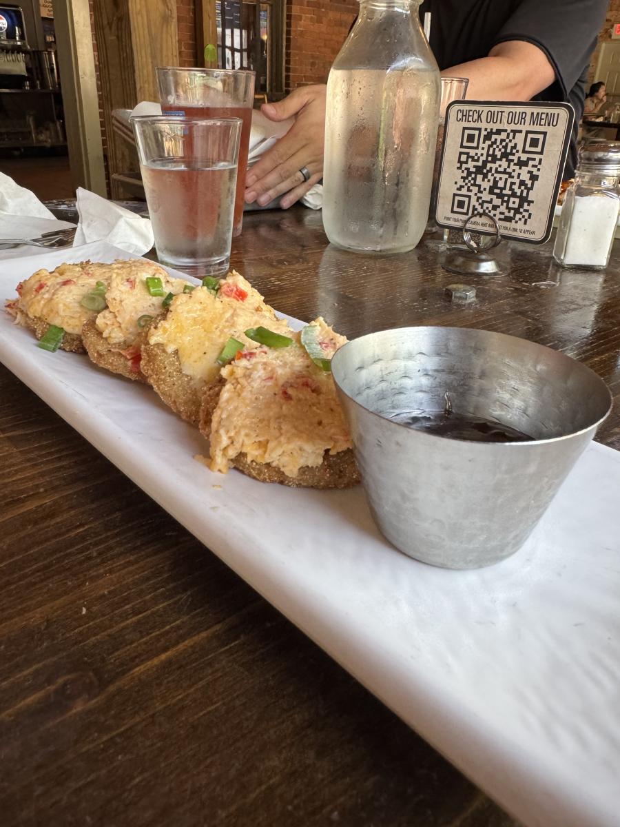 Four fried green tomatoes with a pimento cheese topping on a white marble slab dish at Old Black Bear Brewery. A small tin of dipping sauces sits beside the tomatoes. Someone has a beer and a glass of water behind the appetizer.