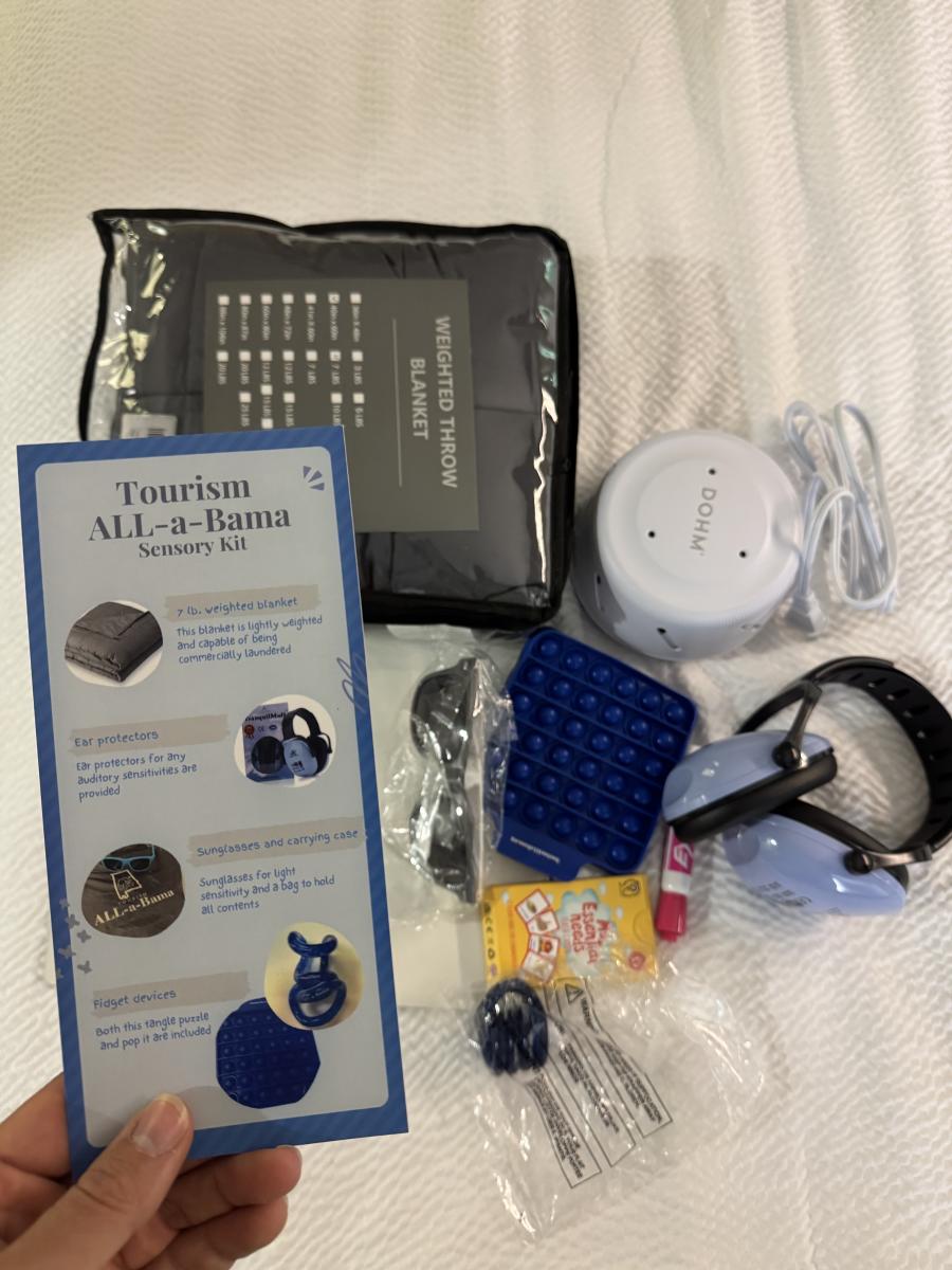 The photo shows a Tourism ALL-a-Bama Sensory Kit laid out on a bed. Items include a gray 7 lb weighted throw blanket in its packaging, a white Dohm white-noise machine with a power cord, blue noise-reducing headphones, a pair of black sunglasses with a carrying case, two fidget devices (a blue pop-it and a blue tangle puzzle), a packet of Smarties candy, and lip balm. A hand is holding the instruction card that describes the kit’s contents and uses.