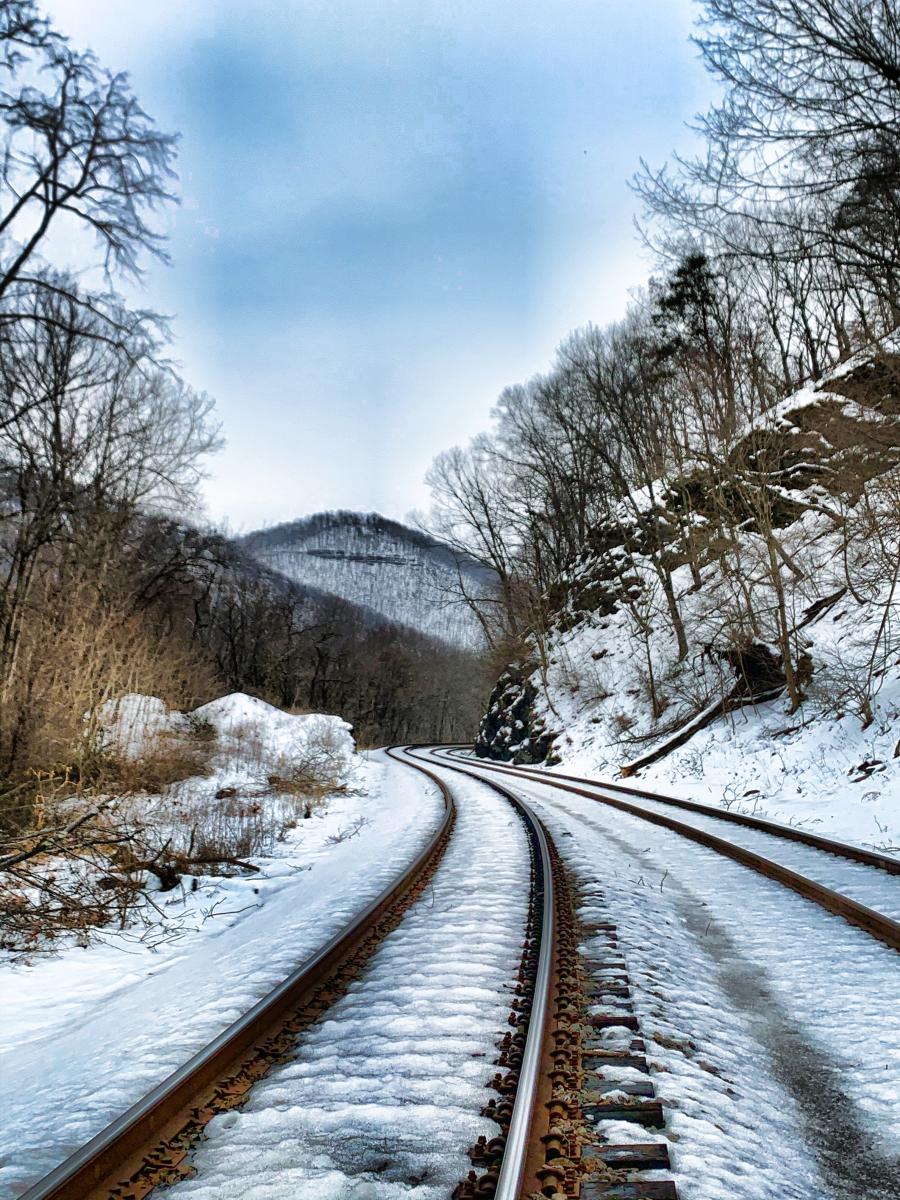 Snow-covered railroad tracks curve through a wooded mountain landscape in Cresaptown, Maryland, with bare winter trees and rolling Appalachian hills in the distance under a cloudy sky.