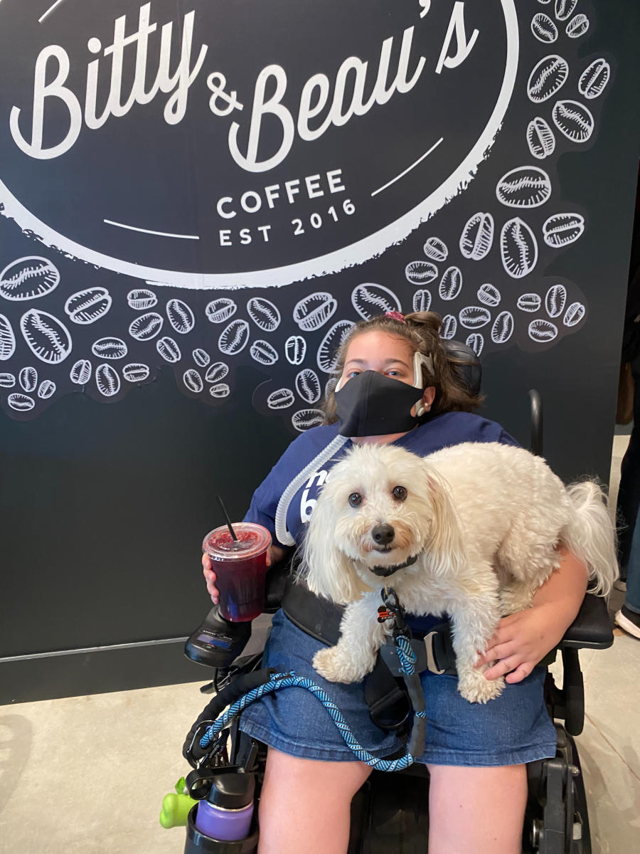 photo shows a woman in a wheelchair, wearing a mask, holding a small white dog in front of a Bitty & Beau's Coffee sign.