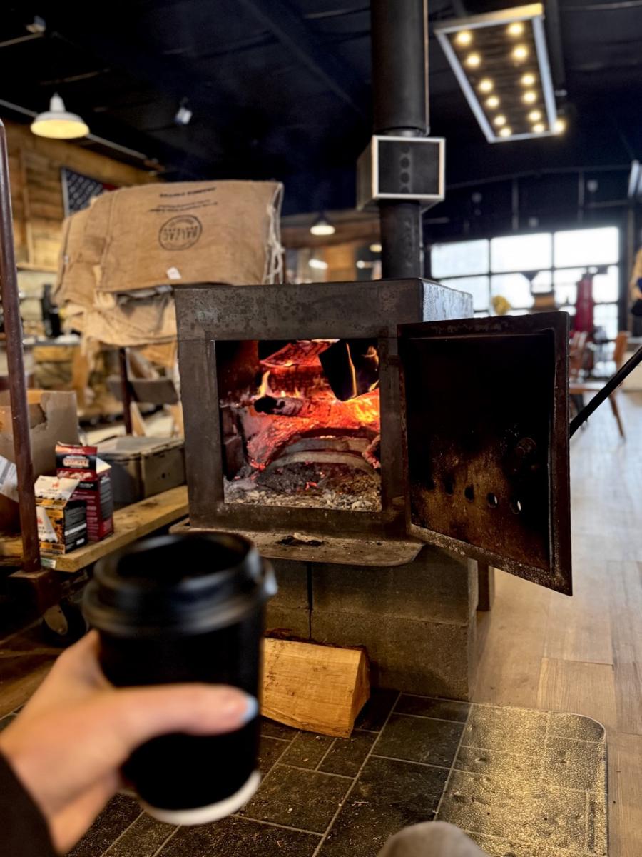 A wood-burning fireplace rests open to show a burning fire, with a hand holding a coffee cup in the foreground and a coffee roastery with wooden floors and a large window in the background.