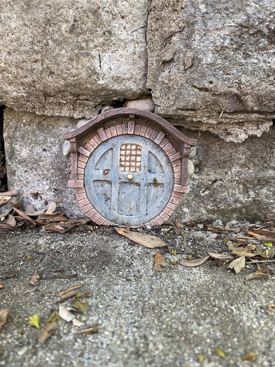 Image is of a small, round, blue fairy door that is along the side walk, built into the retaining wall.