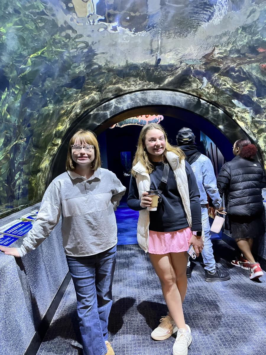 Image is of two teenage girls standing int he tunnel that is covered in glass where the aquarium surrounds them.