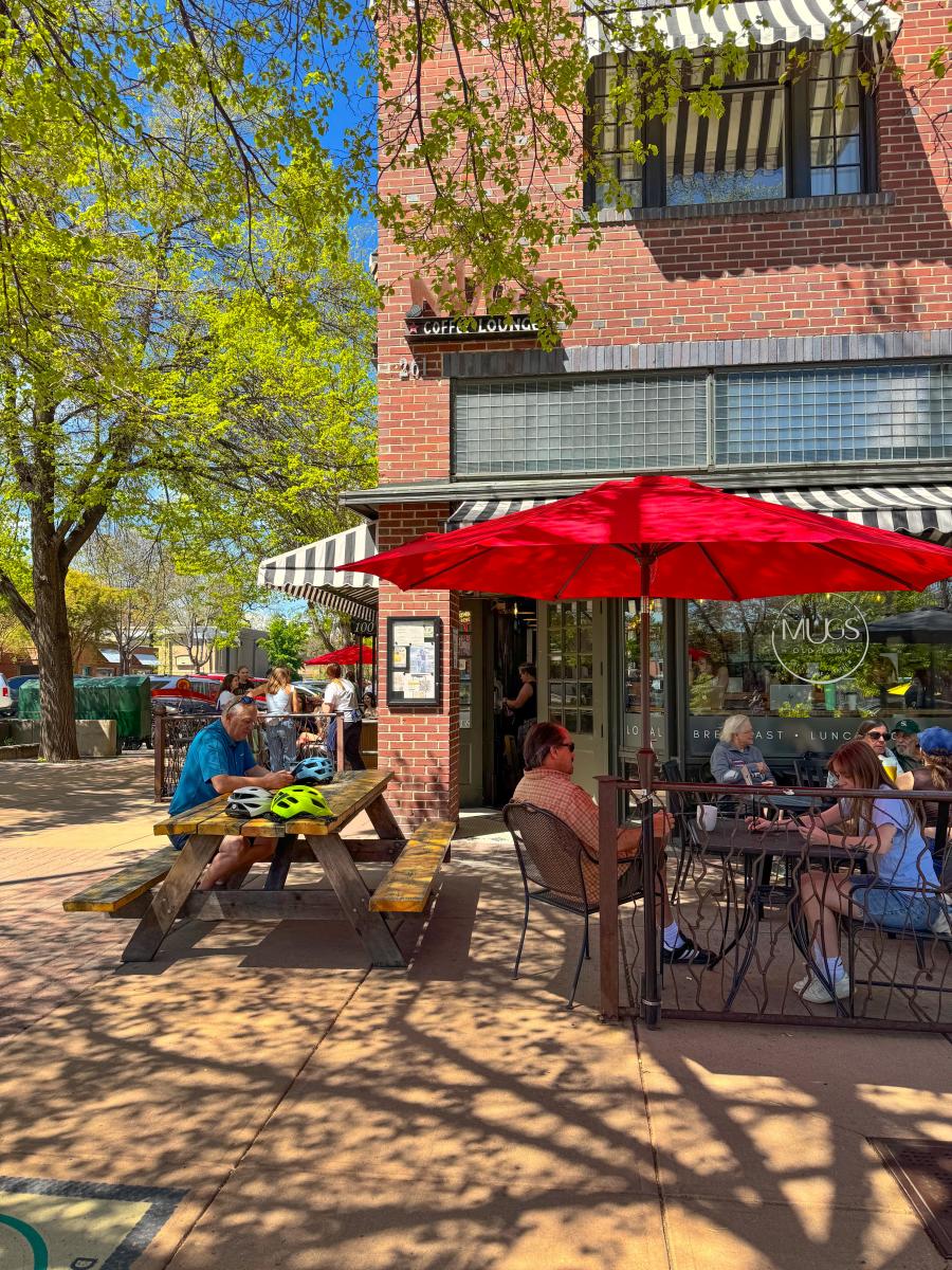 Outside of coffee shop with people sitting at tables underneath red umbrella.