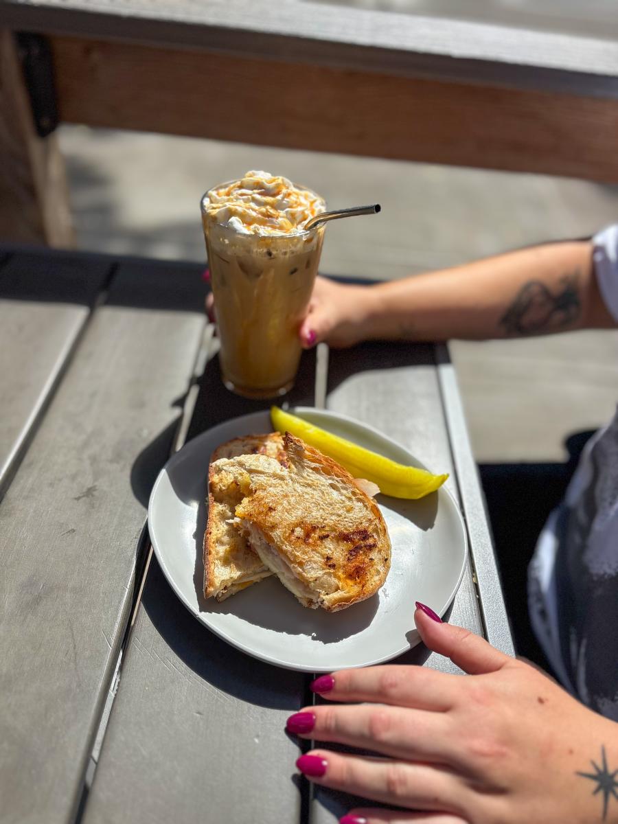 A toasted sandwich with a pickle spear and a coffee drink in a glass on a table.